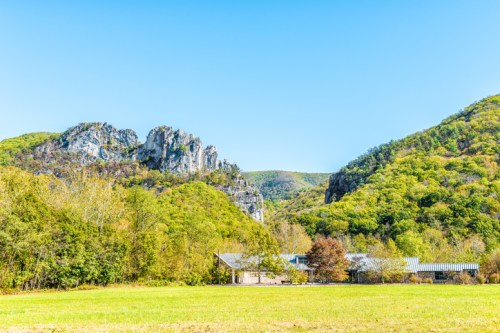 A scenic view of green hills and rocky cliffs under a clear blue sky, with a small building in the foreground.