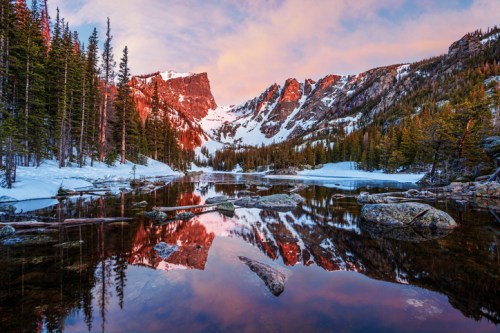 A serene mountain landscape at sunset, with snow-capped peaks reflecting in a calm lake surrounded by pine trees.