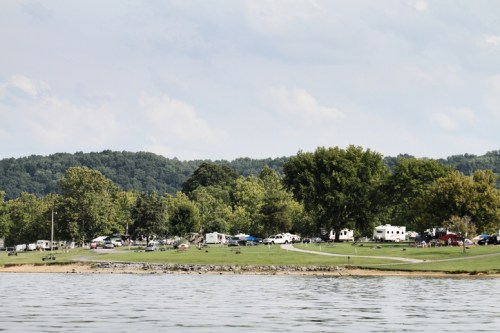 A serene lakeside view of a campground with RVs and trees, set against a backdrop of rolling green hills.