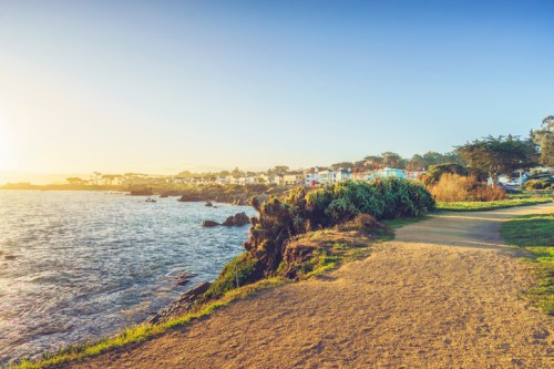 Coastal view with a sandy path, rocky shoreline, and colorful buildings in the distance under a clear sky.