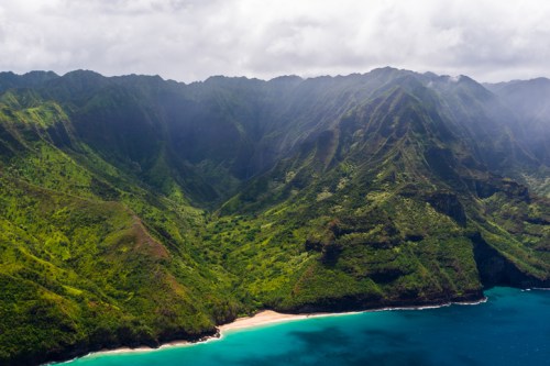 Aerial view of lush green mountains meeting a sandy beach and turquoise ocean under a cloudy sky.