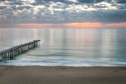 A serene beach scene at sunset, featuring a long pier extending into calm waters under a cloudy sky.