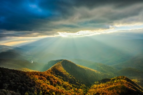 Sunlight breaks through clouds over rolling hills, highlighting autumn foliage and a winding river in the valley below.