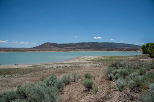 A serene lake surrounded by dry land and shrubs, with mountains in the background under a clear blue sky.