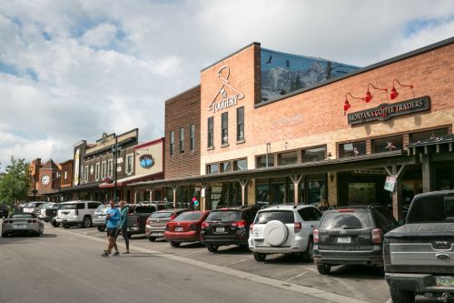A street view of a charming town with shops, parked cars, and people walking under a partly cloudy sky.