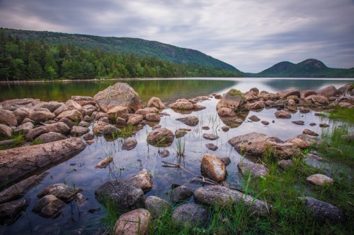 A serene lake surrounded by rocky shores and lush green hills under a cloudy sky.