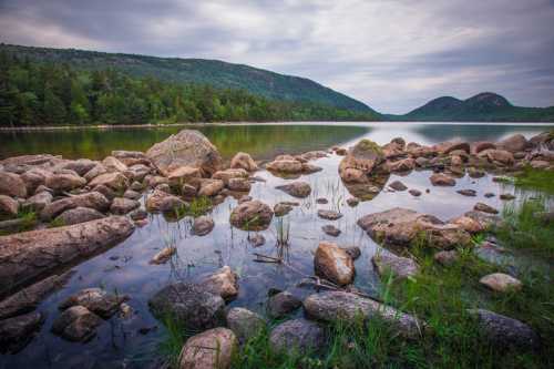 A serene lake surrounded by rocky shores and lush green hills under a cloudy sky.