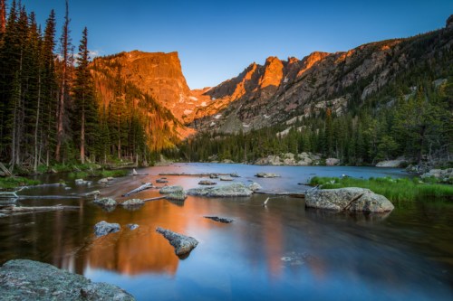 A serene mountain lake at sunset, surrounded by trees and rocky formations, reflecting vibrant colors in the water.