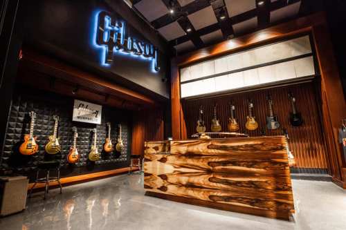 Interior of a guitar shop featuring a wooden counter and a wall displaying various guitars. The Gibson logo is illuminated.