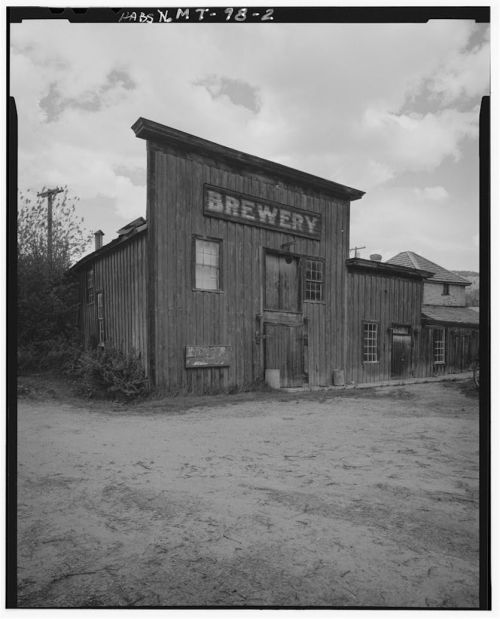 Historic wooden brewery building with a sign, surrounded by dirt and trees under a cloudy sky.
