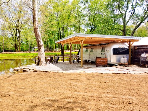 A cozy trailer under a wooden shelter by a pond, surrounded by trees and gravel seating area.