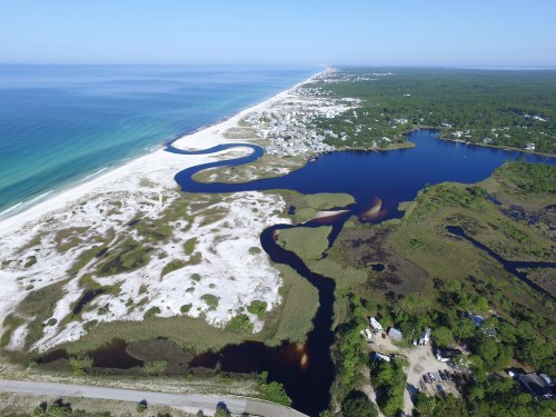 Aerial view of a coastal landscape featuring sandy beaches, winding rivers, and lush greenery under a clear blue sky.