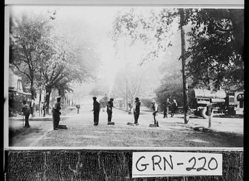 Historic black-and-white photo of soldiers standing on a street, with trees and buildings in the background.