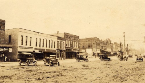 Historic sepia-toned photo of a dusty street lined with early 20th-century cars and storefronts.