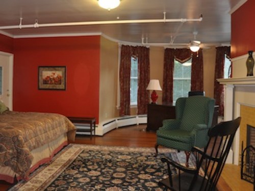 Cozy bedroom with red walls, a large bed, green armchair, desk, and patterned rug. Natural light from windows.