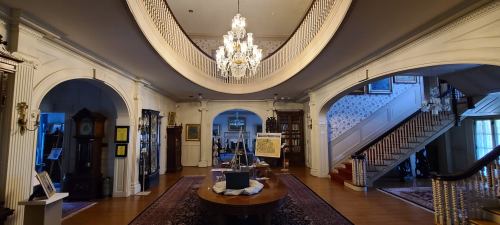 A spacious, elegantly decorated foyer with a chandelier, arched doorways, and a staircase, featuring antique furnishings.