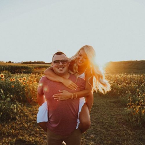 A couple smiles joyfully in a sunflower field at sunset, with the sun glowing behind them.