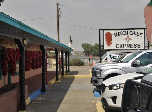 A person stands on a roof near a sign for Hatch Chile Express, with parked cars and a clear sky in the background.