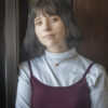 A person with short hair wearing a blue turtleneck and a burgundy top, standing by a window with a thoughtful expression.