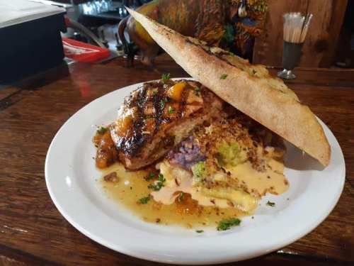 A grilled chicken dish topped with sauce, served on a plate with colorful sides and a large piece of flatbread.