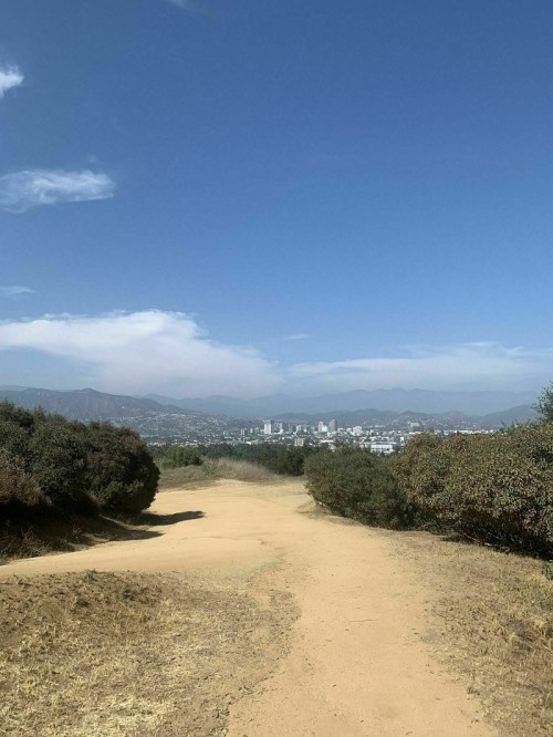 A dirt path leads through greenery, with a city skyline and mountains visible under a clear blue sky.