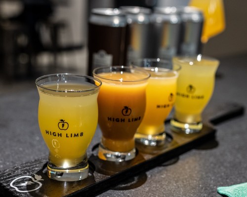 A flight of four colorful drinks in branded glasses on a wooden paddle, with cans in the background.