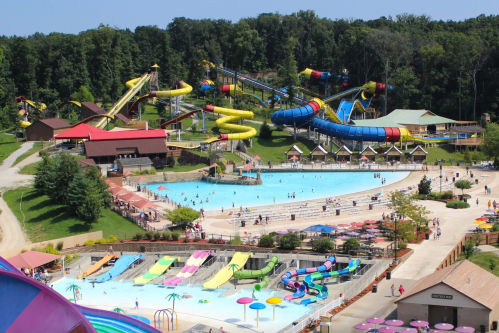 Aerial view of a water park featuring slides, a large pool, and colorful lounge areas surrounded by trees.