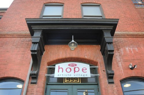 Sign for "Hope Artiste Village" on a brick building, featuring a decorative entrance and large windows.
