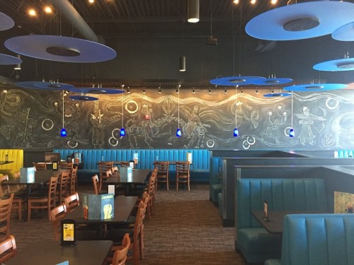 Interior of a restaurant featuring blue pendant lights, a chalkboard mural, and teal booths with wooden tables.