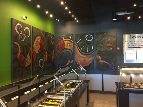 Colorful abstract murals on a green wall above a food serving area with metal trays and a menu board.