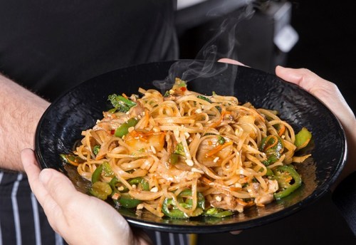 A steaming plate of stir-fried noodles with vegetables, held by two hands against a dark background.