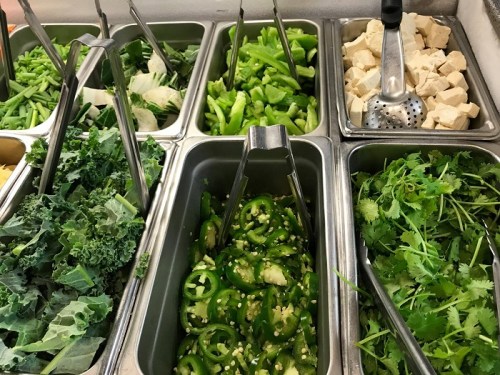 A variety of fresh vegetables and herbs in metal containers, including greens, peppers, and tofu.