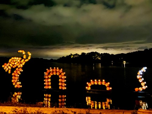 Illuminated pumpkin sculptures reflect on a dark lake under a cloudy night sky.