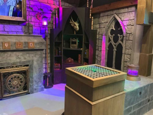 A fantasy-themed room with stone walls, a decorative fireplace, and a wooden table with a grid pattern, illuminated by colorful lights.