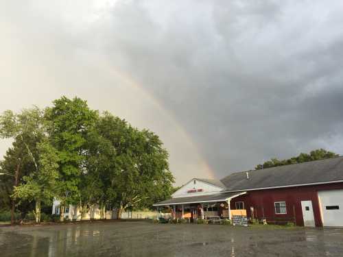 A rainbow arcs over a farm building surrounded by trees under a cloudy sky after rain.