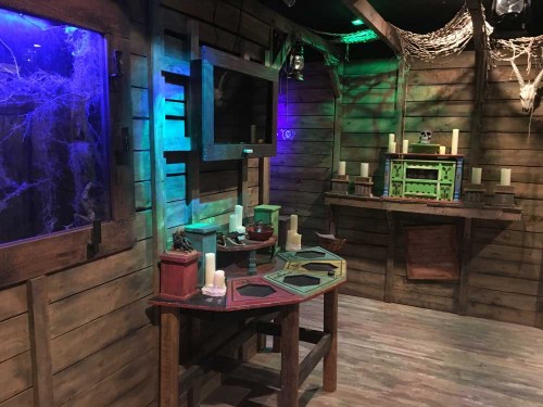 A dimly lit room with wooden walls, a table with colorful boxes, candles, and eerie decorations like cobwebs and a skull.