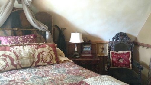 Cozy bedroom with a decorative bed, ornate chair, lamp, and vintage decor against a sloped ceiling.