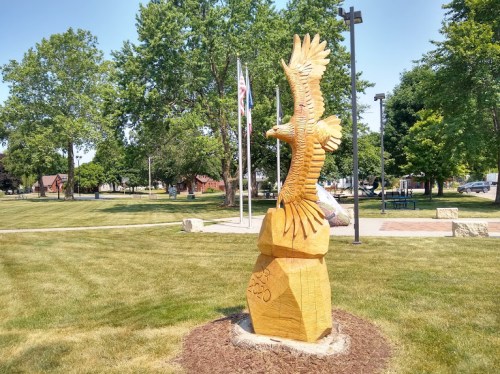Wooden eagle sculpture on a stone base in a park, surrounded by grass and trees, with flags in the background.