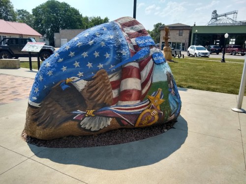 A large painted rock featuring an eagle, American flag, and military medals, set in a public area with greenery.