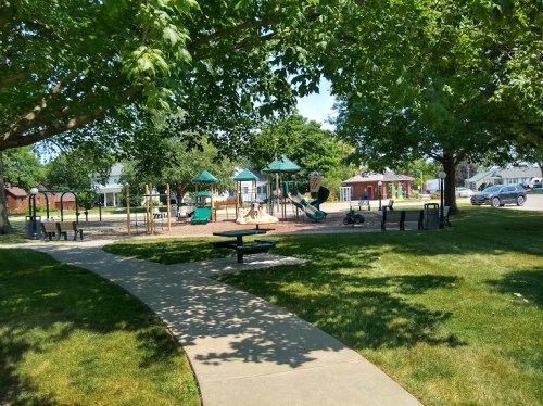 A sunny park with a playground, picnic tables, and a winding path surrounded by trees and green grass.