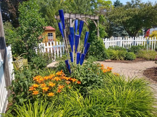 A vibrant garden featuring orange lilies, lush greenery, and a striking blue glass sculpture amidst a white picket fence.