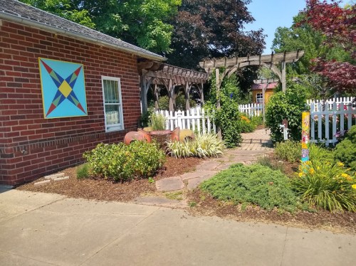 A colorful quilt square adorns a brick wall beside a garden with a white picket fence and a trellis.