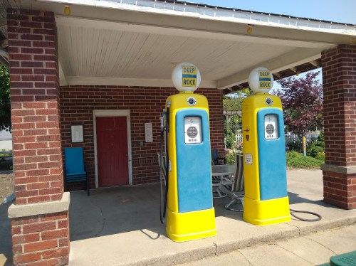 Two vintage gas pumps in bright yellow and blue, located under a brick shelter with a red door in the background.