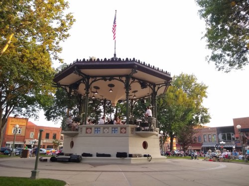 A bandstand in a park with musicians performing, surrounded by trees and a small crowd enjoying the outdoor concert.