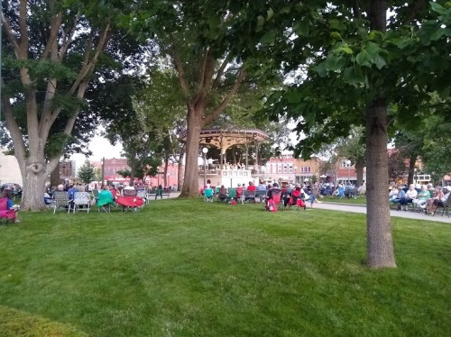 A park scene with people sitting on the grass and in chairs, gathered around a gazebo surrounded by trees.