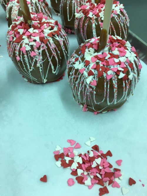 Chocolate-covered apples decorated with pink, white, and red heart-shaped sprinkles on a white surface.