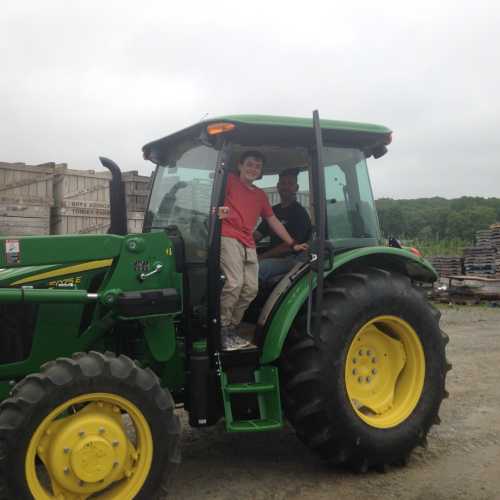 A child and an adult sit in a green tractor, surrounded by crates and a rural landscape.