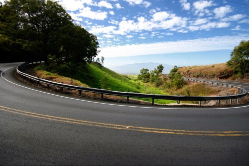 A winding road curves through lush green hills under a blue sky with scattered clouds.