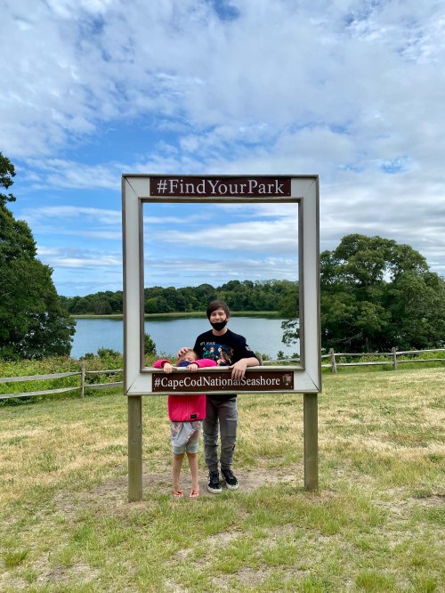 Two children stand inside a park photo frame with a lake and trees in the background at Cape Cod National Seashore.