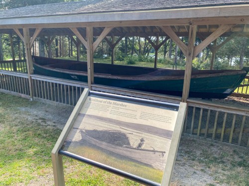 A wooden boat displayed under a shelter, with an informational sign in front about its history.